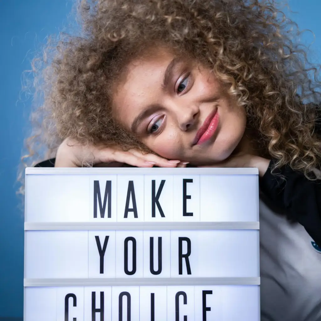 Smiling woman with curly hair posing with a lightbox sign that reads 'Make Your Choice' against a blue background.