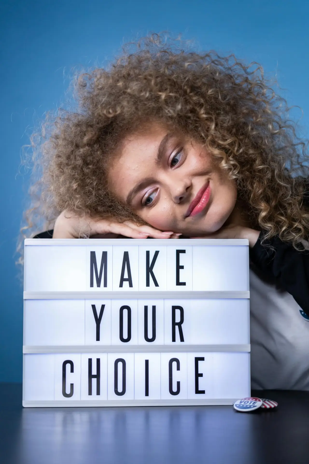 Smiling woman with curly hair posing with a lightbox sign that reads 'Make Your Choice' against a blue background.