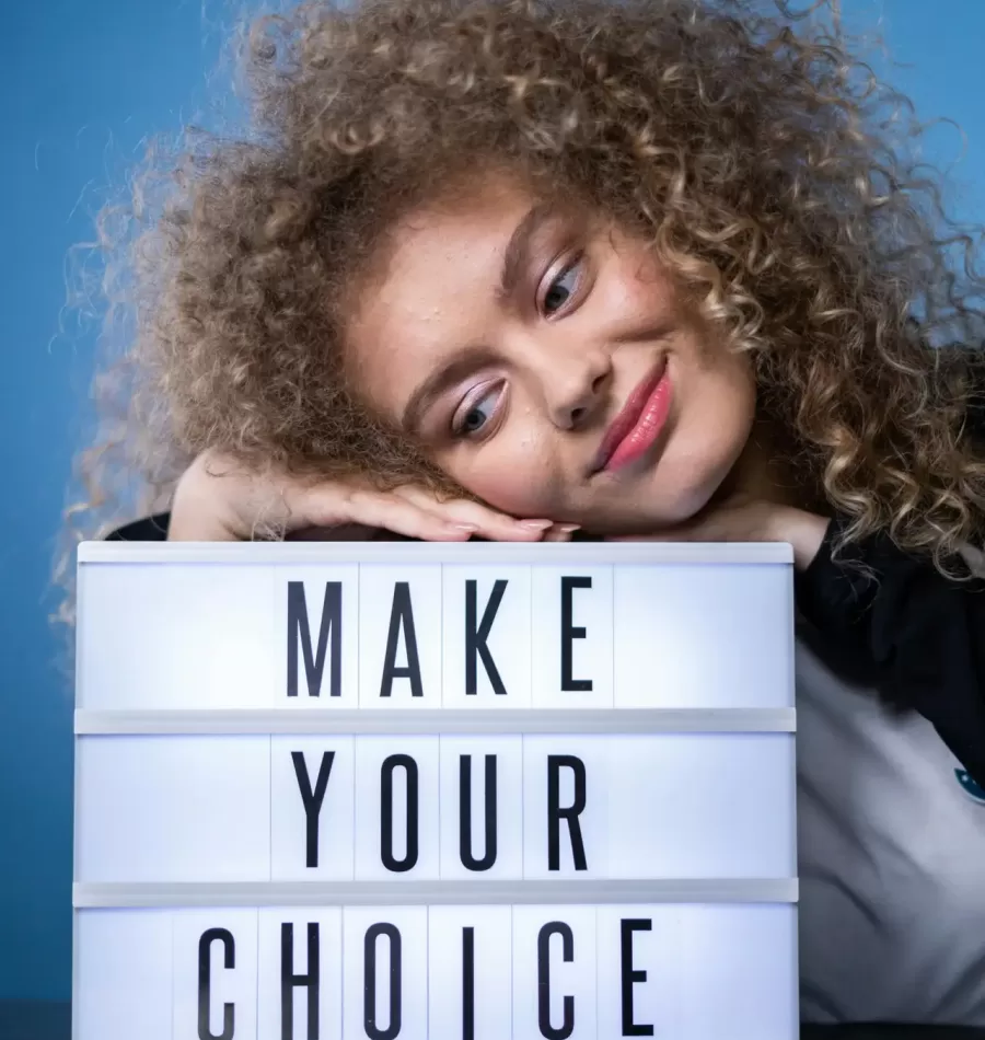 Smiling woman with curly hair posing with a lightbox sign that reads 'Make Your Choice' against a blue background.